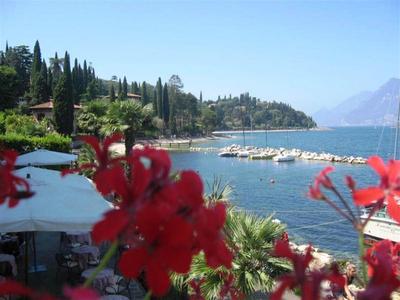 Vista su un lago con barche a vela, palme e fiori rossi in primo piano sotto un cielo limpido.