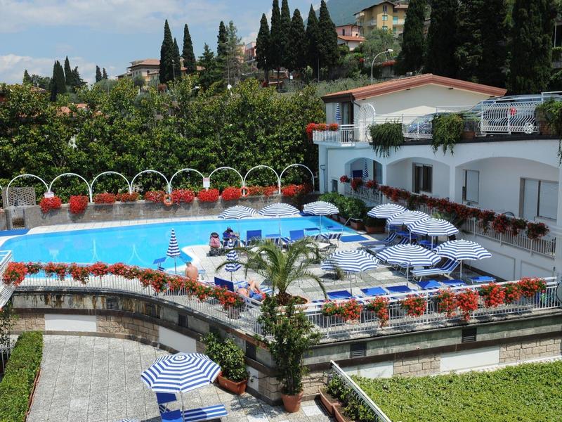 Piscina all'aperto con lettini e ombrelloni accanto a un edificio bianco dell'hotel in un ambiente mediterraneo.