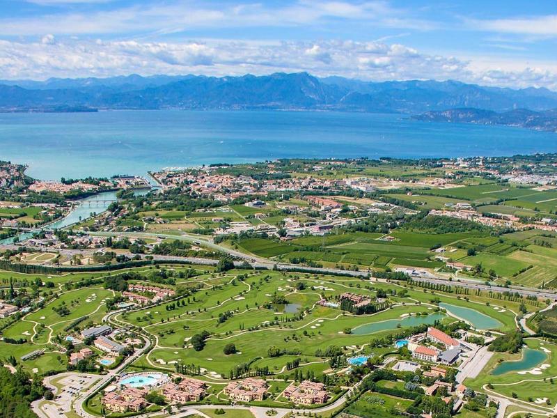 Aerial view of green landscape with golf course and lake in the background under blue sky.