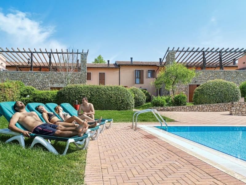 People relax on sun loungers beside an outdoor pool in front of a hotel with pergolas.