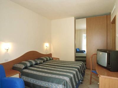 Prosto urządzony pokój hotelowy z podwójnym łóżkiem, szafą z lustrem i małym telewizorem.