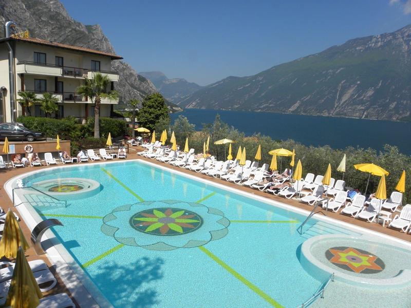 Basen hotelowy z leżakami i parasolami, w tle widok na góry i jezioro.