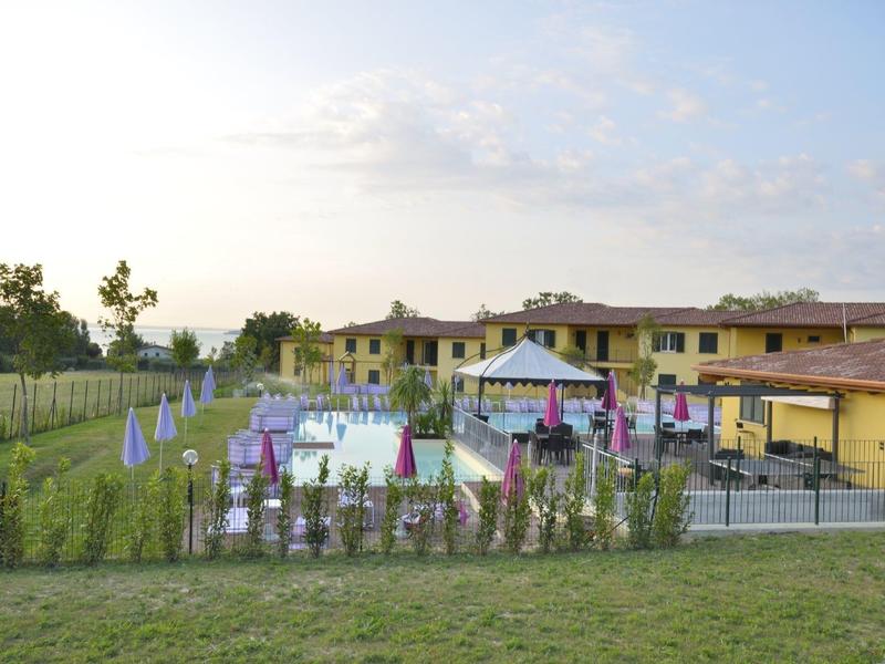 Großer Garten mit Zelt, Stühlen und Tischen vor gelbem Gebäude unter blauem Himmel.