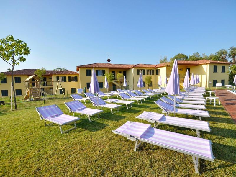 Mehrere weiße Liegestühle mit blauen Polstern stehen auf grünem Rasen vor gelbem Gebäude bei blauem Himmel.