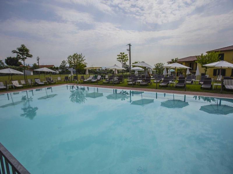 Großer klarer Pool mit Liegestühlen und Sonnenschirmen in ruhiger Gartenanlage unter bewölktem Himmel.