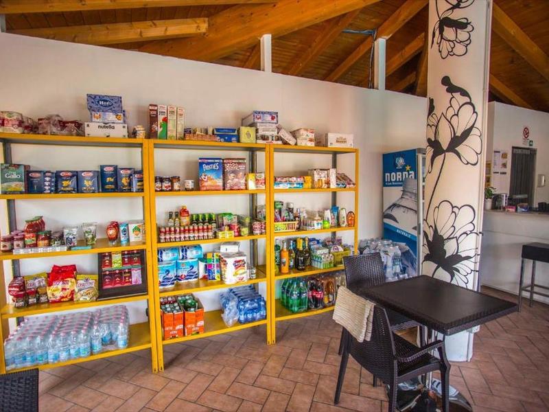 Kleiner Laden mit gelben Regalen, gefüllt mit Lebensmitteln und Küchenutensilien in hellem Raum.