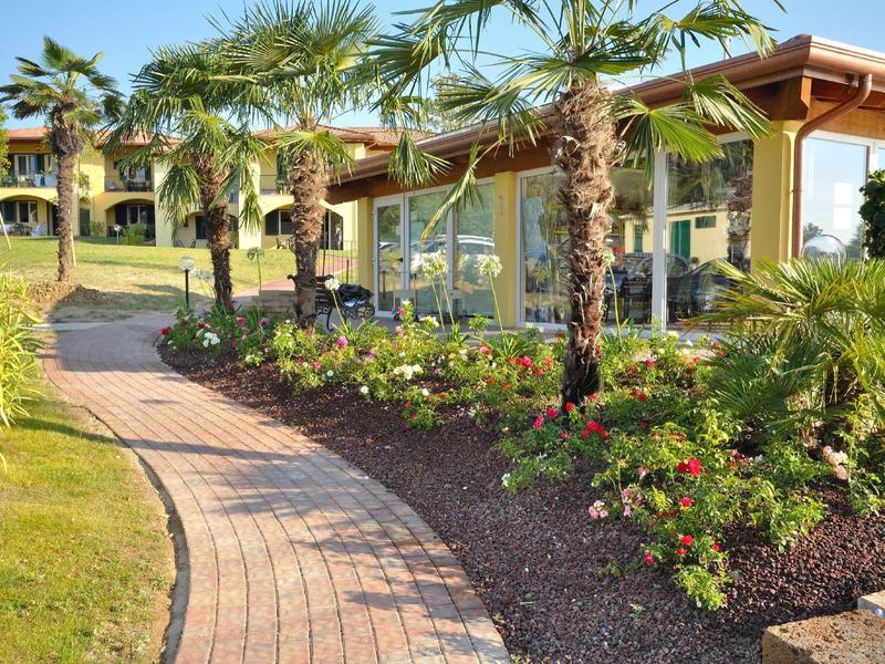 Gepflasterter Weg führt an Palmen und bunten Blumen am Gebäude im sonnigen Garten vorbei.