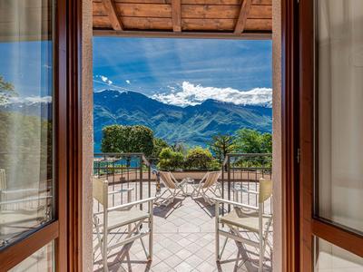 Balkon mit weißen Stühlen und Blick auf grüne Bäume und hohe Berge unter blauem Himmel.