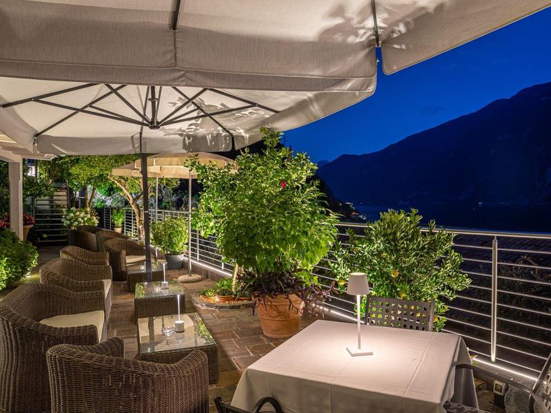 Gemütliche Terrasse mit Tischen, Stühlen, Pflanzen und großer Sonnenschirmbeleuchtung am Abend.