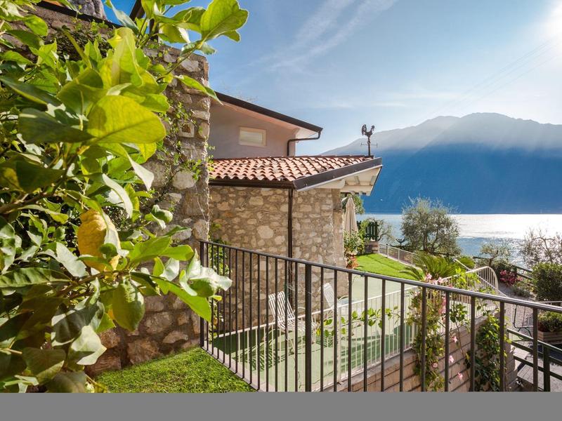 Haus mit Steinmauer, grünem Garten, Metallgeländer und Bergsee im Hintergrund unter blauem Himmel.