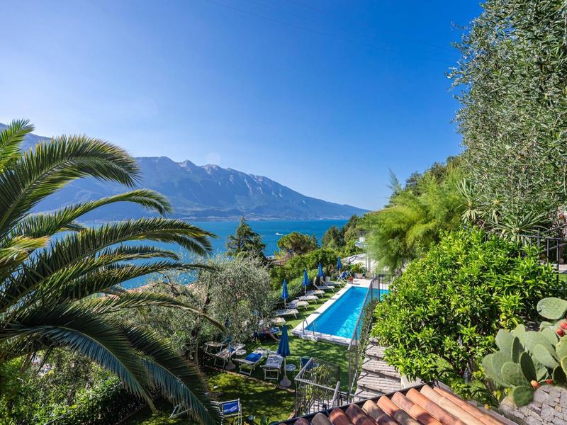 Blick auf Pool mit Liegen, umgeben von Palmen und Büchern, im Hintergrund Berge unter blauem Himmel.