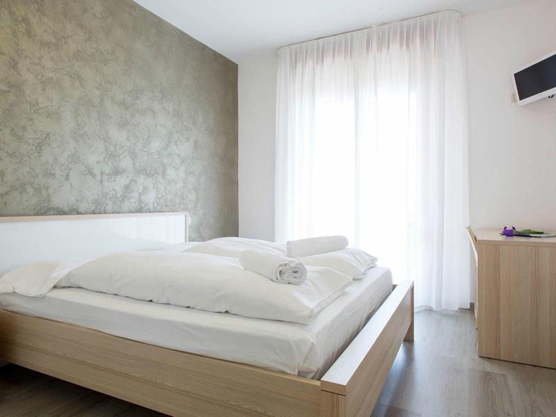 Modernes Schlafzimmer mit großem Bett, hellen Vorhängen und minimalistischen Holzmöbeln.