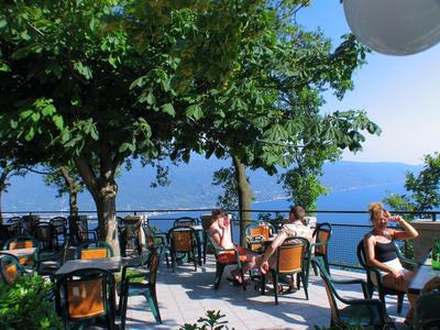 Freiluft-Café mit grünen Stühlen und Tischen, Gästen, Bäumen und Blick aufs blaue Meer.