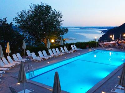 Beleuchteter Pool mit Liegestühlen und Sonnenschirmen, dahinter Meer und Hügel im Abendlicht.