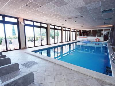 Innen-Schwimmbad mit blauem Wasser, bodentiefen Fenstern und grauen Sitzstühlen am Beckenrand.