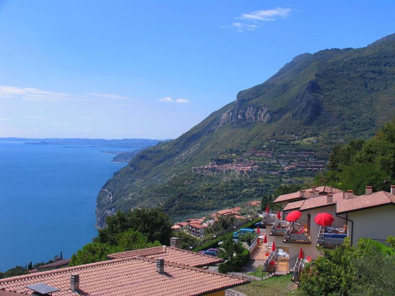 Berge mit grüner Vegetation, Häuser mit roten Dächern und blauer See unter klarem Himmel.