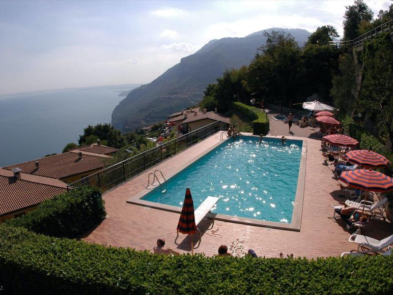 Schwimmbad mit blauen Wasser, umgeben von Sitzplätzen mit roten Sonnenschirmen und Berglandschaft im Hintergrund.
