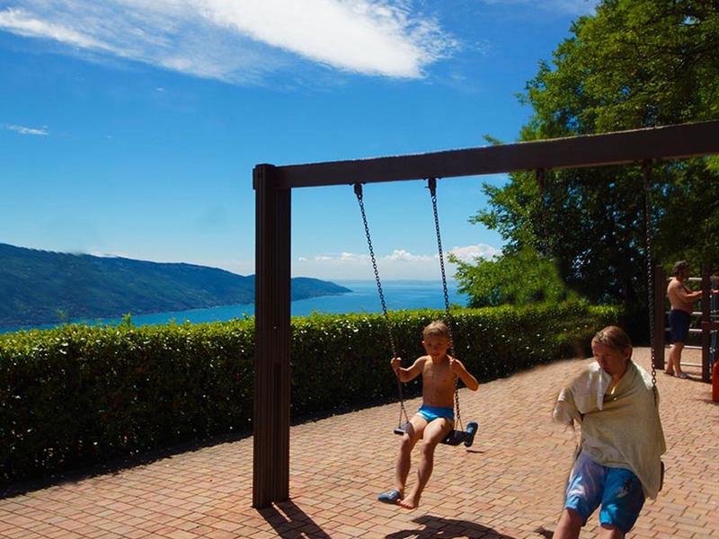 Kinder schaukeln auf einer Holzschaukel vor einer grünen Hecke mit Bergen und blauem Himmel.