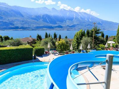 Schwimmbad mit blauem Wasserrutschenpool, grünen Büschen und Bergen im Hintergrund bei Sonnenschein.