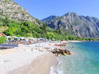Strand mit Kieselsteinen, türkisfarbenem Wasser, Bergen im Hintergrund und Sonnenschirmen am Ufer.