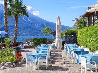 Blaue Stühle und weiße Tische auf sonniger Terrasse mit Blick auf See, Palmen und Berge.