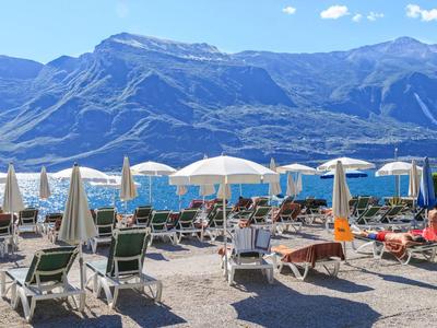 Sonnige Terrasse am See mit Liegestühlen und weißen Sonnenschirmen vor Bergkulisse.
