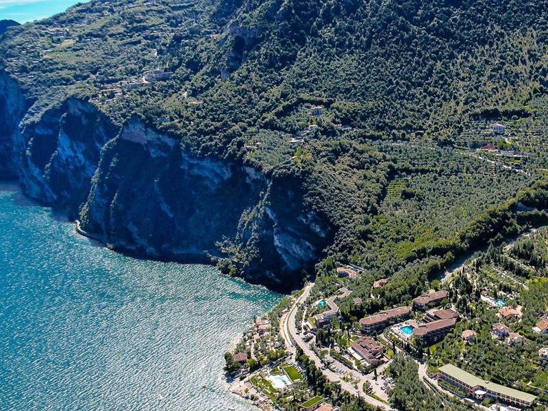 Steile grüne Küstenklippe trifft auf klares blaues Meer mit kleinen Gebäuden am Ufer.