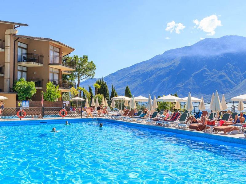 Großer blauer Pool vor Hotel mit Bergen im Hintergrund und Sonnenschirmen am Beckenrand.