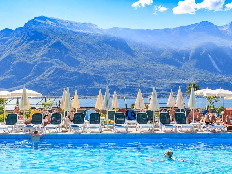 Ein Schwimmbad mit blauen Liegestühlen und weißen Sonnenschirmen vor hohen Bergen unter blauem Himmel.