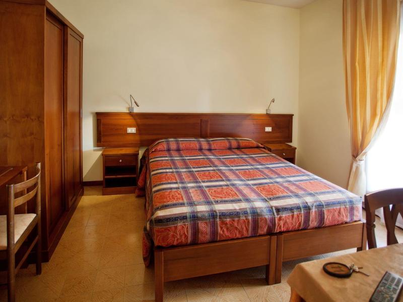 Schlicht eingerichtetes Schlafzimmer mit Doppelbett, Holzmöbeln und großem Fenster mit gelben Vorhängen.