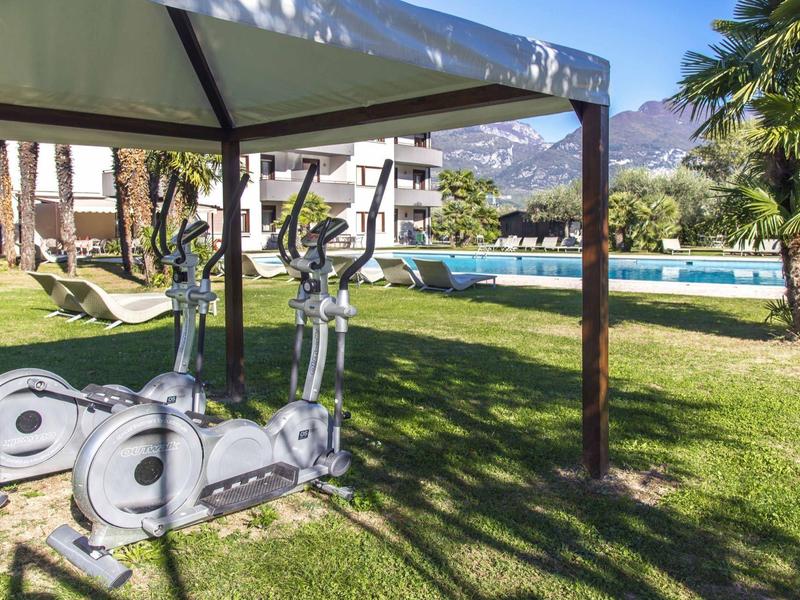 Outdoor-Fitnessgeräte und Liegestuhl stehen auf grünem Rasen vor einem Pool unter blauem Himmel.