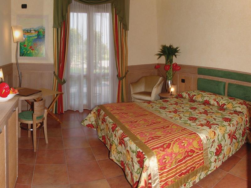 Bright hotel room with two beds, desk, chair, and balcony with curtains.