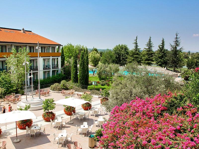 Terrazza dell'hotel con tavoli, sedie, piante in fiore e vista su pini sotto un cielo sereno.