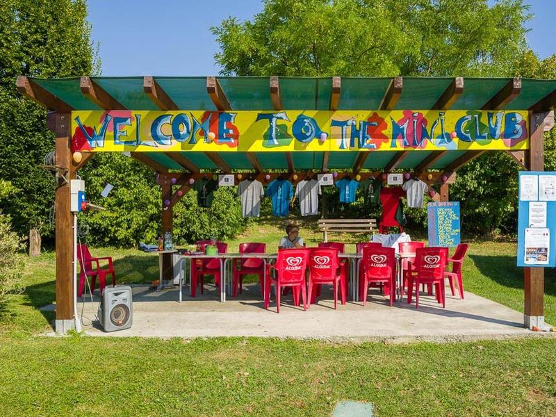 Bar sulla spiaggia colorata con sedie rosse e pergolato verde sotto cielo azzurro e alberi.