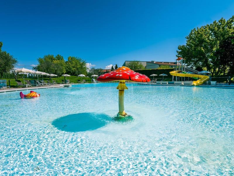 Grande piscina all'aperto con giochi d'acqua e scivolo sotto un cielo azzurro.