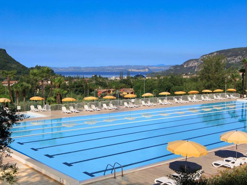 Großer Außenpool mit blauen Bahnen, umgeben von Liegestühlen und gelben Sonnenschirmen, unter blauem Himmel.
