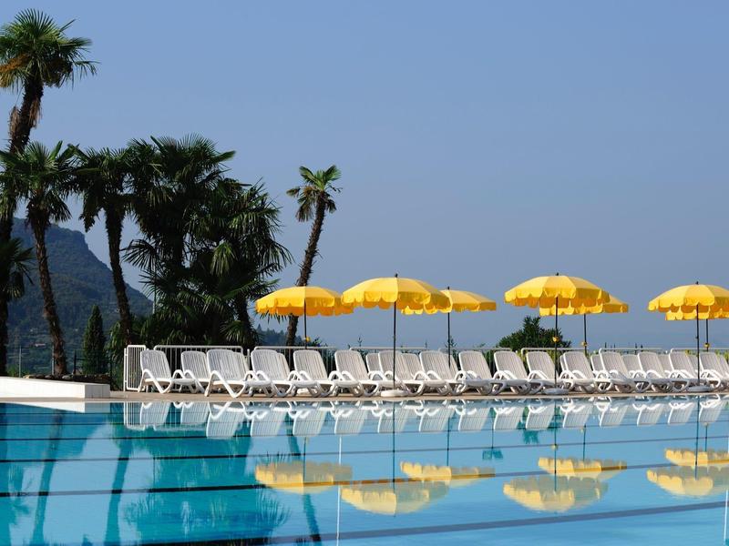 Pool mit klar blauem Wasser, weißen Liegestühlen und gelben Sonnenschirmen, Palmen im Hintergrund.