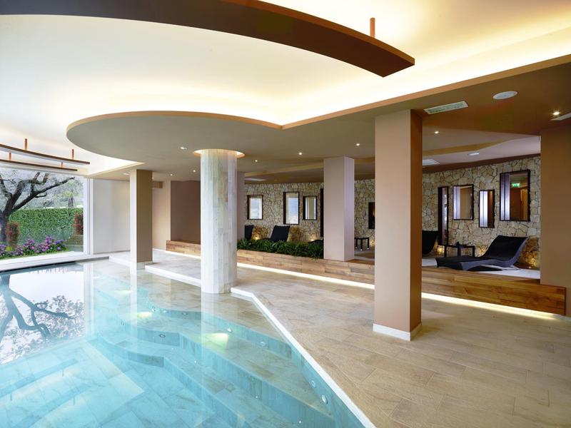 Moderner Innenpool mit hellem Deck, Säulen und Relaxbereich mit Sofas in warmen Tönen.