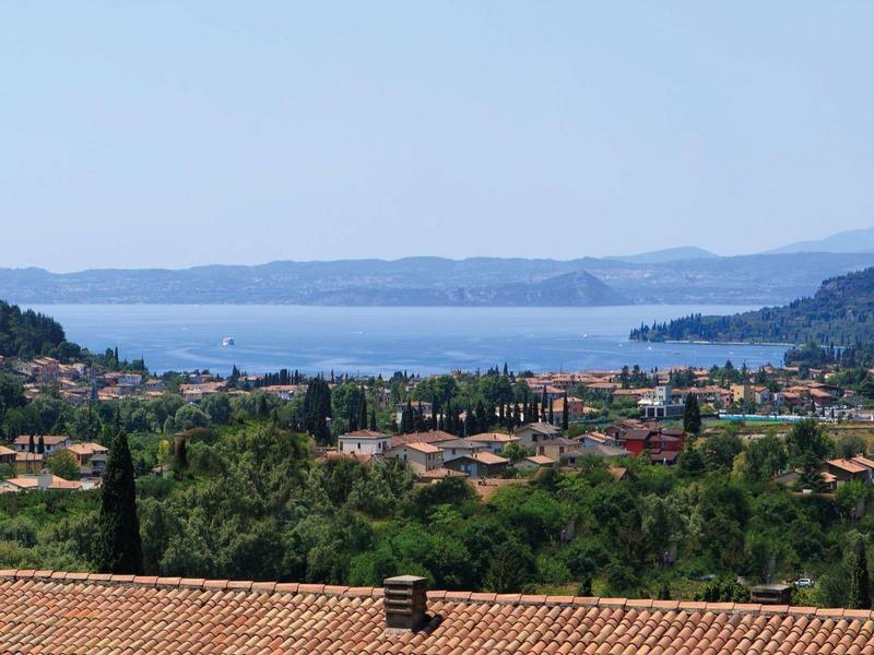 Landschaft mit Häusern, grünen Hügeln und weiter Sicht auf blauen See unter klarem Himmel.