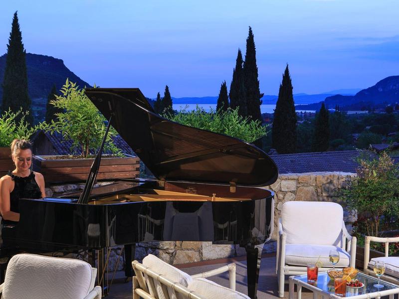 Frau spielt Flügelklavier auf Terrasse mit weißen Stühlen und mediterraner Landschaft im Hintergrund.