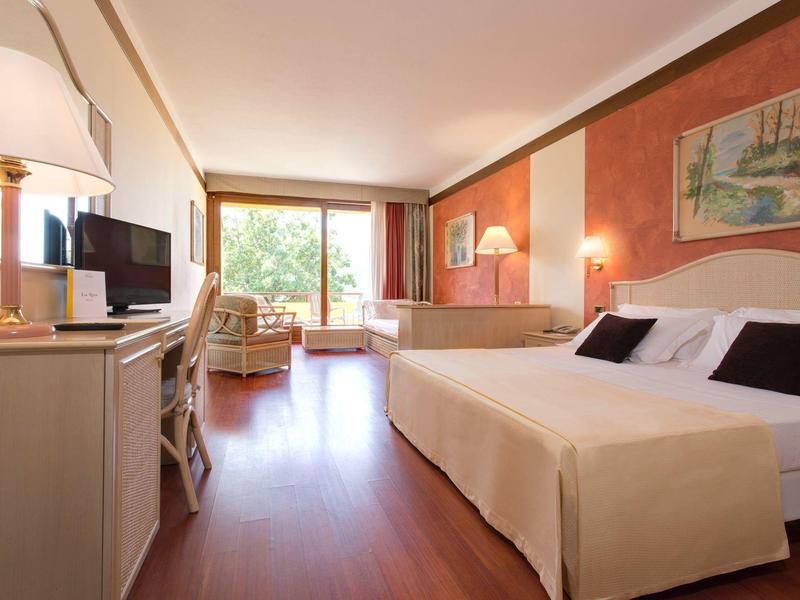 Helles Hotelzimmer mit großem Fenster, Doppelbett, Fernseher und Sitzbereich mit Stühlen und Tisch.