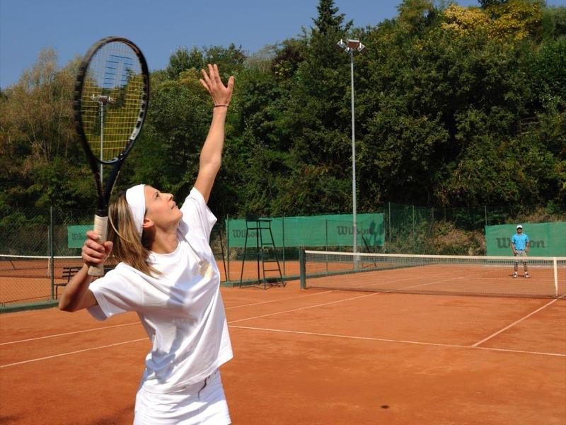 Frau in weißer Sportkleidung spielt Tennis auf rotem Sandplatz bei klarem Himmel.