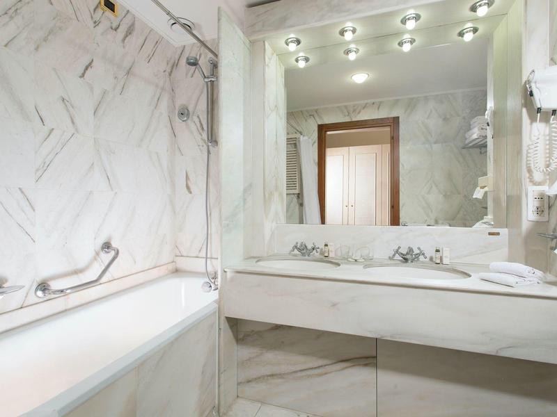 Helles Badezimmer mit Marmorwänden, großer Spiegel, Doppelwaschbecken und Badewanne mit Haltegriff.