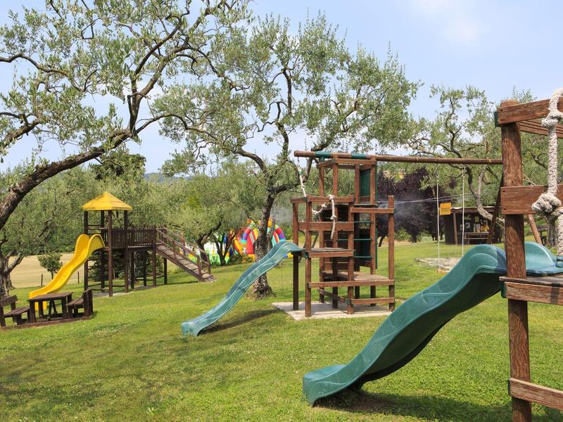 Kinderspielplatz mit Rutschen, Klettergerüsten und Bäumen auf grüner Wiese unter blauem Himmel.