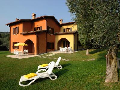 Zweistöckiges Haus in warmen Farben mit Garten, Sonnenliegen und großem Baum im Vordergrund.