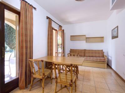 Heller Raum mit Holztisch, drei Holzstühlen, Klappcouch, Fliesenboden und großem Fenster mit braunen Vorhängen.