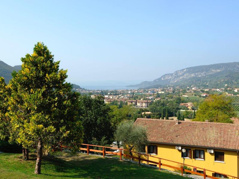 Landschaft mit grünem Baum, gelbem Haus und Bergen im Hintergrund unter klarem Himmel.