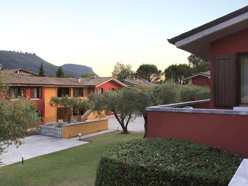 Rotes Wohnhaus mit Garten, Büschen und Bäumen vor hügeliger Landschaft bei Sonnenaufgang.