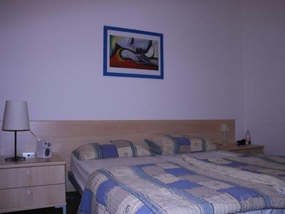 Sypialnia z podwójnym łóżkiem, wzorzystą narzutą, stolikiem nocnym, lampką i obrazem na ścianie.