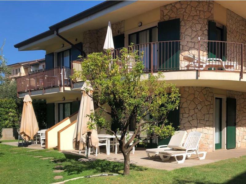 Hotel dwupiętrowy z balkonami, ogrodem, parasolami i leżakami na trawniku.